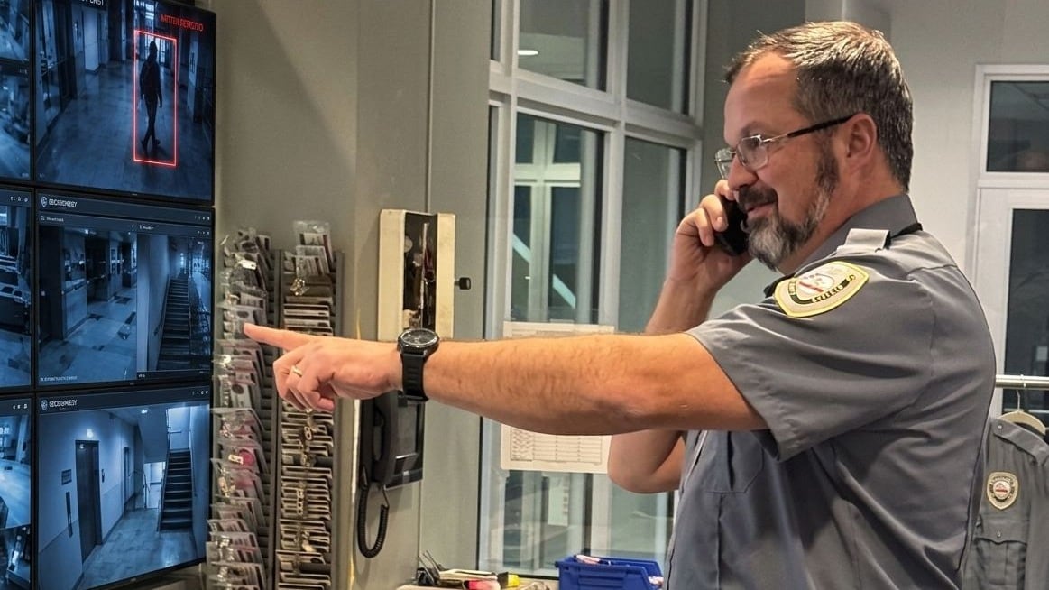 Securemedy officer in a grey uniform on the phone, pointing to surveillance monitors