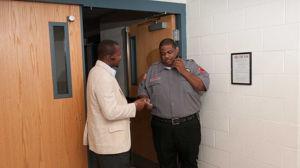 Securemedy officer in a grey uniform on a radio call while assisting a visitor at a doorway